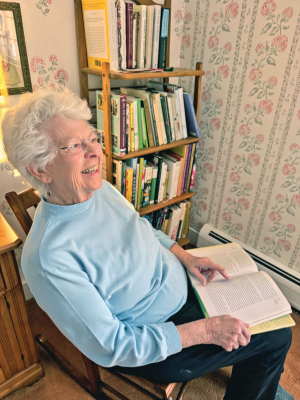 Photo by Scooter MacMillan.
Joan Weed sits with a gardening book in front of a shelf filled with her gardening books.