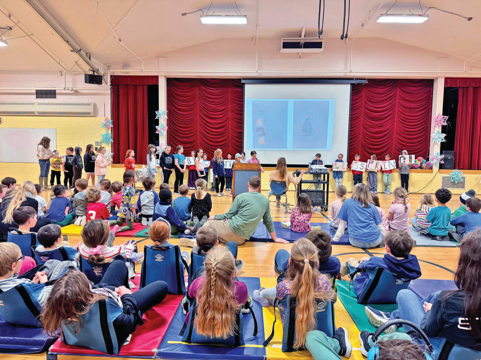 Courtesy photo. In April, third graders lead an all-school assembly, as each grade level does during the school year. Because of technical limitations like inadequate lighting, these student presentations take place on the floor in front of the stage.