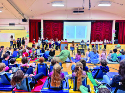 Courtesy photo. In April, third graders lead an all-school assembly, as each grade level does during the school year. Because of technical limitations like inadequate lighting, these student presentations take place on the floor in front of the stage.